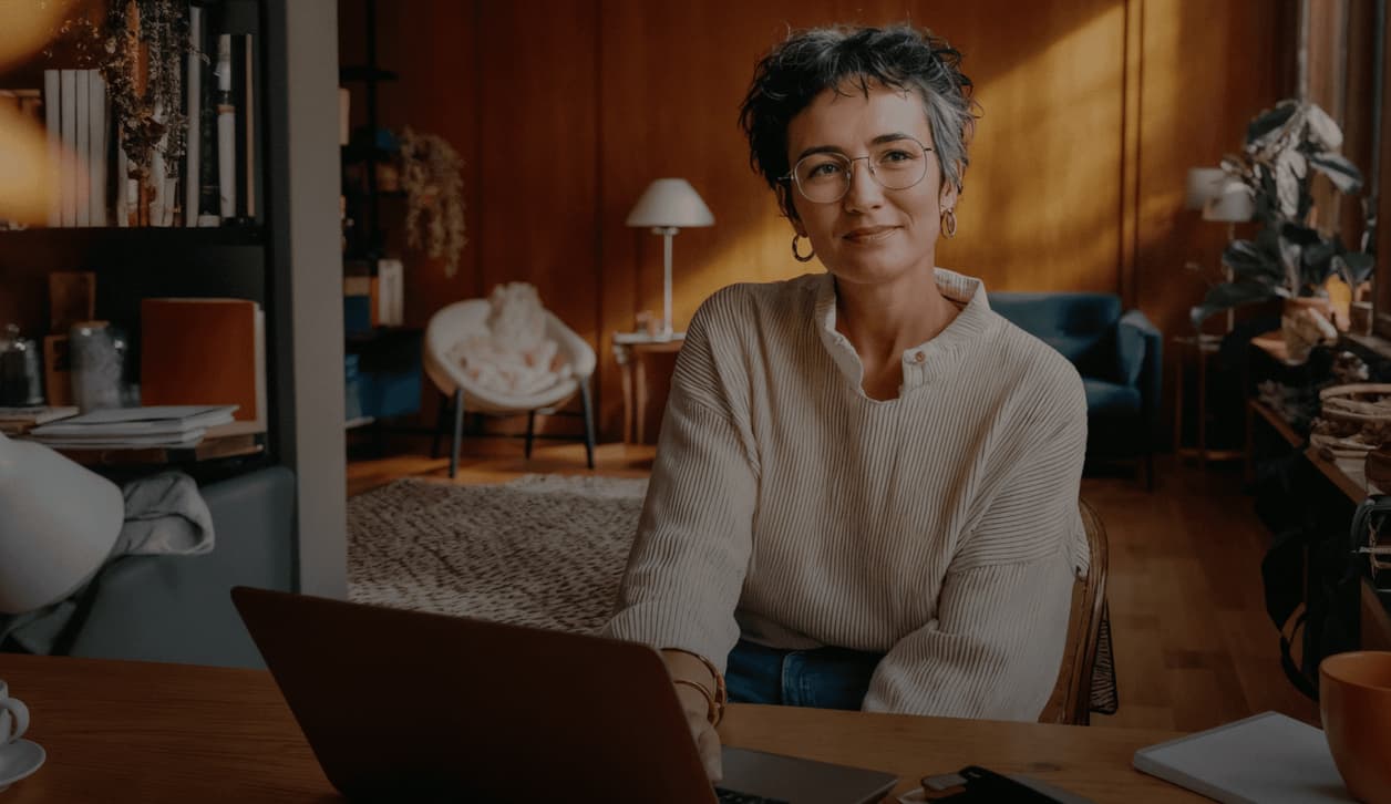 A woman with short hair and glasses sits confidently at a desk in a cozy well-decorated room