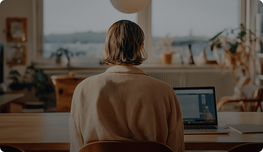 A person with short hair is sitting at a desk in a softly lit room working on a laptop